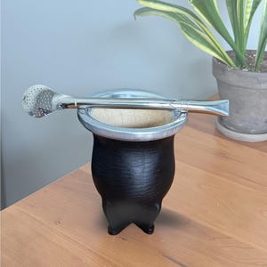 May include: A black mate gourd with a smooth, rounded body and a silver-colored metal rim, resting on three small feet. A long, silver-colored metal bombilla straw with a perforated, spoon-shaped filter end is placed horizontally across the top rim.