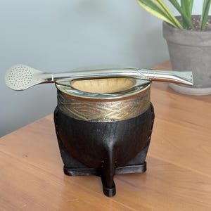 May include: A dark brown leather-wrapped mate gourd with a metallic rim featuring an etched geometric pattern. A silver-toned metal bombilla with a perforated, flat, oval-shaped filter end rests horizontally across the top of the gourd opening.