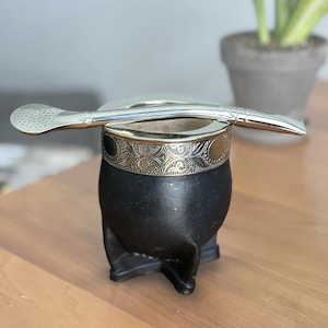 May include: A matte black mate gourd with a wide, silver-toned metal rim featuring an intricate embossed floral pattern. A long, polished silver-toned metal bombilla straw with a perforated filter end rests horizontally across the top of the gourd.