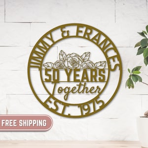 Può includere: Un'opera d'arte da parete in metallo color oro. Il design circolare presenta i nomi "Jimmy & Frances", le parole "50 Years Together" e l'anno "EST. 1975". Un motivo floreale è centrato sopra le parole "50 Years".