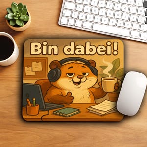 May include: A mousepad featuring a cartoon beaver wearing headphones and holding a coffee cup. The beaver is sitting at a desk with a laptop, notepad, and the German phrase "Bin dabei!" printed on the mousepad. A white mouse and keyboard are in the background.
