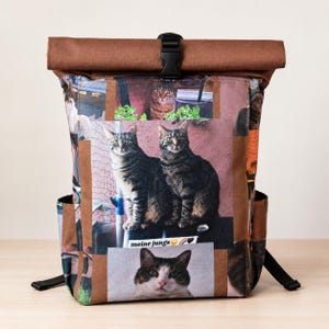 Op de afbeelding: Een roll-top rugzak met een collage-ontwerp van katten. De tas heeft een bruine bovenkant en randen, een zwarte gesp en zijvakken. De tekst "meine jungs" is zichtbaar op de voorkant. Afmetingen niet beschikbaar.