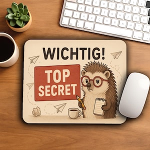 Könnte beinhalten: Beige Mauspad mit der Aufschrift "WICHTIG! TOP SECRET" und einem Cartoon-Igel mit Brille. Der Igel hält einen Bleistift und einen Notizblock. Eine weiße Maus, eine Tastatur und eine kleine Sukkulentenpflanze sind ebenfalls sichtbar.
