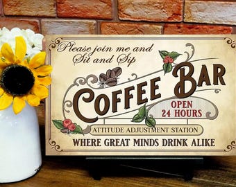 May include: A vintage-style sign with the words "Coffee Bar" in brown lettering, decorated with coffee beans and leaves. The sign reads "Open 24 Hours" and "Attitude Adjustment Station". A vase with sunflowers is in the foreground.