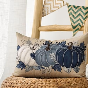 May include: A rectangular beige throw pillow featuring a printed illustration of three pumpkins in shades of white, light blue, and dark navy blue, surrounded by dark blue and white leaves, resting on a woven textured seat with a wooden frame.