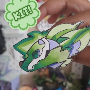 May include: A paper cutout of a stylized green and white creature head with purple accents and dark outlines. A light green scalloped speech bubble graphic with the text KIT! is positioned above the creature head illustration.
