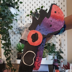 Puede incluir: Una máscara de papel hecha a mano con una base negra y un patrón vibrante inspirado en una galaxia en rojo, morado y azul. La máscara incluye un detalle circular rojo en el ojo, un contorno blanco en forma de alubia y bordes dentados superpuestos.