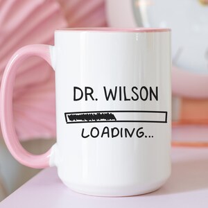 May include: White ceramic mug with a pink handle and rim. The mug features the text "DR. WILSON" above a loading bar graphic, with "LOADING..." below. The mug is set against a soft pink background.