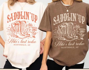 May include: Two t-shirts, one ivory and one espresso, each with a graphic design. The design includes the text "SADDLIN' UP" above a drawing of cowboy boots, flowers, and a skull. Below the graphic is "Niki's last rodeo, Scottsdale, AZ."