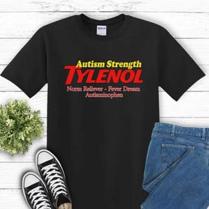 Könnte beinhalten: Schwarzes T-Shirt mit dem Text "Autism Strength TYLENOL" in Gelb und Rot. Unter dem Wort "TYLENOL" stehen die Worte "Norm Reliever - Fever Dream Autisminophen" in Rot. Das Shirt wird mit blauen Jeans und schwarz-weißen Sneakers kombiniert.