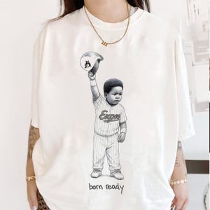 Peut inclure: T-shirt crème avec une illustration en noir et blanc d'un jeune joueur de baseball tenant une casquette. Le joueur porte un uniforme à rayures avec le mot "Expos" et le texte "born ready" en bas.