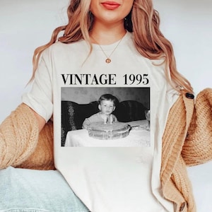 K&ouml;nnte beinhalten: Cremefarbenes T-Shirt mit einem Schwarz-Wei&szlig;-Foto eines Kindes mit Geburtstagskuchen. Der Text "VINTAGE 1995" ist in fetter, schwarzer Schrift &uuml;ber dem Foto aufgedruckt. Die Person tr&auml;gt eine goldene Halskette und eine beige Strickjacke.