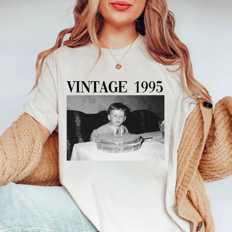 K&ouml;nnte beinhalten: Cremefarbenes T-Shirt mit einem Schwarz-Wei&szlig;-Foto eines Kindes mit Geburtstagskuchen. Der Text "VINTAGE 1995" ist in fetter, schwarzer Schrift &uuml;ber dem Foto aufgedruckt. Die Person tr&auml;gt eine goldene Halskette und eine beige Strickjacke.