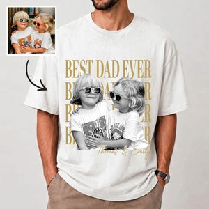 May include: Cream-colored t-shirt featuring a black and white photo of two children wearing sunglasses. The words "BEST DAD EVER" are printed in gold, along with the names "Thomas & Emily". The shirt is a casual, oversized style.