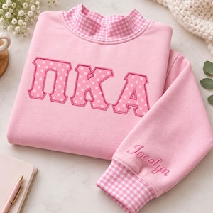 Peut inclure: Un sweat-shirt rose clair avec un col montant et des poignets à motif vichy rose et blanc. Le devant présente les lettres grecques Pi Kappa Alpha en rose à pois blancs. Le prénom Jocelyn est brodé sur la manche du vêtement.