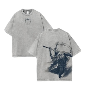 以下が含まれることがあります： 2枚のグレーのTシャツ。Tシャツの前面にはシンボルが付いた円形のデザインがあります。Tシャツの背面には、杖を持っている人物の暗いグラフィックがあります。Tシャツは半袖で、クルーネックです。