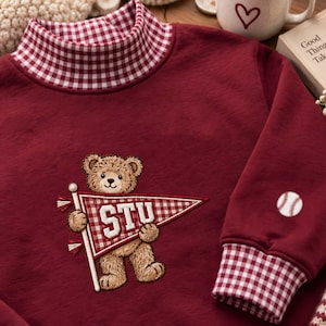 Peut inclure: Un sweat-shirt bordeaux doté d'un col montant à motif vichy rouge et blanc. Au centre, un ours en peluche marron brodé tient un fanion vichy rouge et blanc avec l'inscription STU. Une balle de baseball est brodée sur la manche.