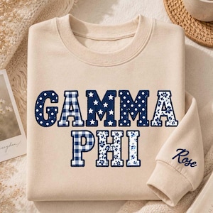May include: A beige crewneck sweatshirt featuring the text GAMMA PHI in large, dark blue patterned letters. The letters display various designs including gingham, stars, polka dots, and floral prints. The name Rose is embroidered on the sleeve.