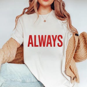Może przedstawiać: Kremowy t-shirt z napisem "ALWAYS" w pogrubionej czerwieni. Koszulka ma swobodny styl, prawdopodobnie wykonana z bawełny. Beżowy kardigan jest zarzucony na ramiona.