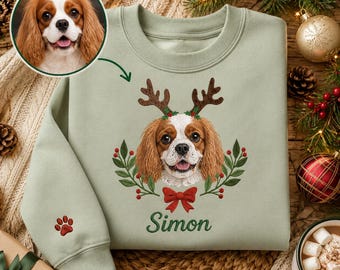 May include: A sage green sweatshirt featuring an embroidered Cavalier King Charles Spaniel wearing reindeer antlers, surrounded by holly leaves and berries. Below the dog is a red bow and the name Simon. A small red paw print is on the sleeve.