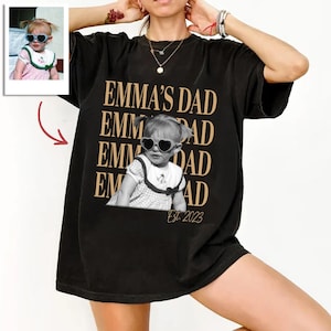 May include: A black short-sleeved t-shirt featuring a repeating tan text graphic that reads EMMA'S DAD. Centered is a black and white photograph of a young child wearing heart-shaped sunglasses and a collared dress. The text Est. 2023 appears below.