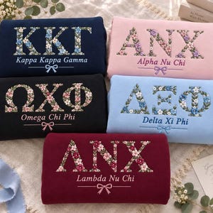 Könnte beinhalten: Fünf gefaltete Sweatshirts in Marineblau, Hellrosa, Schwarz, Hellblau und Kastanienbraun. Jedes zeigt gestickte griechische Buchstaben mit Blumenmustern, den Namen der Organisation und eine kleine Schleifengrafik unter dem Text.