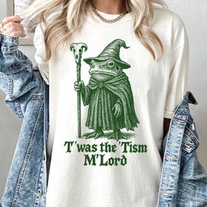Peut inclure: T-shirt blanc cassé avec une illustration verte d'une grenouille sorcier tenant un bâton. La grenouille porte un chapeau pointu et une cape. Le texte "T'was the 'Tism M'Lord" est imprimé en dessous.