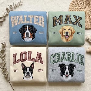 Peut inclure: Quatre sweat-shirts pliés en bleu clair, vert sauge, crème et gris chiné. Chacun présente le portrait d'un chien et un nom dans une police à carreaux : WALTER, MAX, LOLA et CHARLIE, avec EST. 2023 imprimé sous les noms sur chaque vêtement.