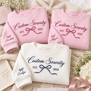 May include: Three crewneck sweatshirts in light pink and white. Two pink sweatshirts feature dark pink embroidery reading Custom Sorority EST. 2026 with a bow, and names Scarlett or Abigail on the sleeve. One white sweatshirt has navy text and bow.
