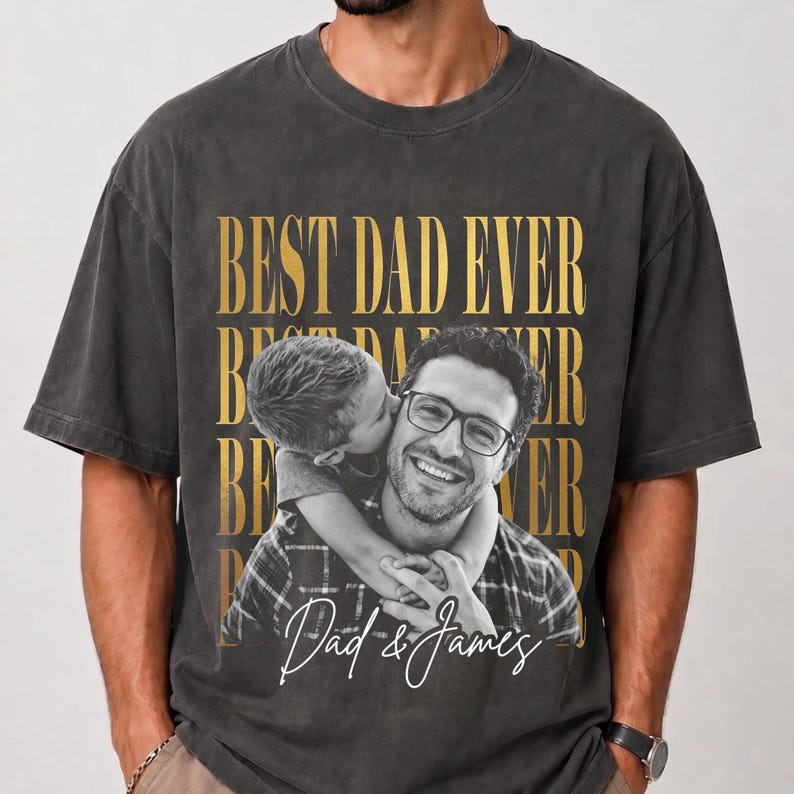K&ouml;nnte beinhalten: Dunkelgraues T-Shirt mit goldenem Aufdruck "BEST DAD EVER" und einem Schwarz-Wei&szlig;-Foto eines Mannes und eines Kindes. Das Foto zeigt ein Kind, das den Mann k&uuml;sst. Der Text "Dad & James" ist in Wei&szlig; geschrieben.