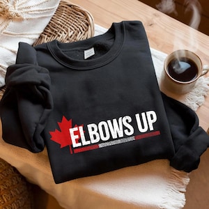 Puede incluir: Sudadera negra con una hoja de arce roja y el texto "Elbows Up" en blanco con una franja roja y blanca debajo.