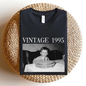 K&ouml;nnte beinhalten: Schwarzes T-Shirt mit dem Text "VINTAGE 1995" &uuml;ber einem Schwarz-Wei&szlig;-Foto eines Kindes mit Geburtstagskuchen. Das T-Shirt ist auf einer gewebten, kreisf&ouml;rmigen Oberfl&auml;che gefaltet.