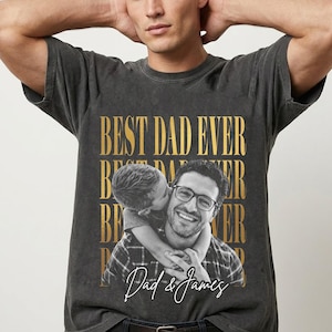 K&ouml;nnte beinhalten: Dunkelgraues T-Shirt mit goldenem Aufdruck "BEST DAD EVER". Das Shirt zeigt ein Foto eines Mannes mit Brille, der von einem Kind gek&uuml;sst wird. Der Text "Dad & James" ist in einer Schreibschrift geschrieben.