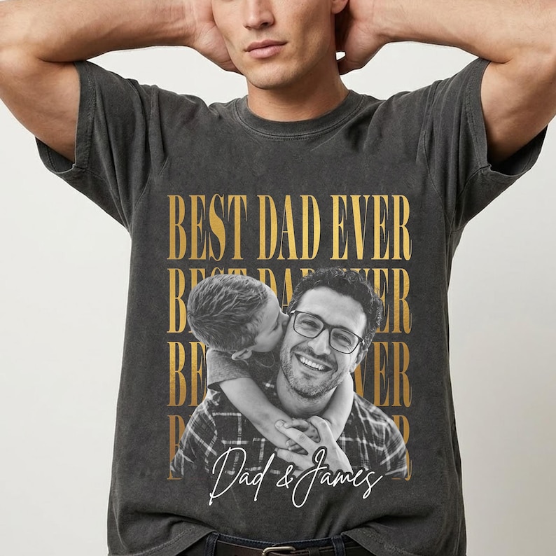K&ouml;nnte beinhalten: Dunkelgraues T-Shirt mit goldenem Aufdruck "BEST DAD EVER". Das Shirt zeigt ein Foto eines Mannes mit Brille, der von einem Kind gek&uuml;sst wird. Der Text "Dad & James" ist in einer Schreibschrift geschrieben.