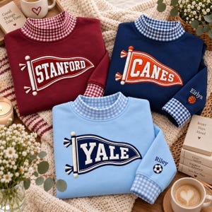 Könnte beinhalten: Drei langärmelige Sweatshirts in Kastanienbraun, Marineblau und Hellblau. Jedes hat einen Stehkragen und Ärmelbündchen mit blau-weißem Vichy-Muster. Gestickte Wimpel zeigen die Schriftzüge STANFORD, CANES und YALE. Die Ärmel sind mit den Namen Eirlys und Riley sowie kleinen Sportball-Symbolen versehen.