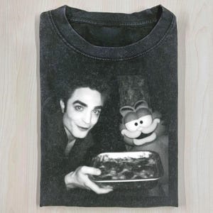 May include: Black and white graphic t-shirt featuring a vintage image of a man and a cartoon cat holding a dish. The t-shirt has a crew neck and short sleeves. The image is centered on the front of the shirt.