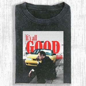 May include: Dark gray t-shirt featuring a graphic with the text "It's all GOOD man" in red. The graphic shows a man in a suit, a yellow car, and a white background. The shirt is folded.