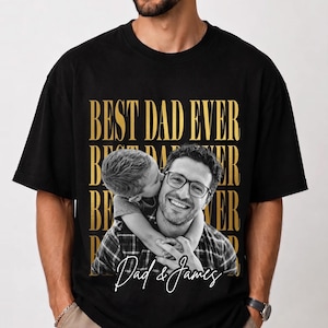 K&ouml;nnte beinhalten: Schwarzes T-Shirt mit goldenem Aufdruck "BEST DAD EVER". Das Shirt zeigt ein Schwarz-Wei&szlig;-Foto eines Mannes mit Brille, der von einem Kind gek&uuml;sst wird. Der Text "Dad & James" ist kursiv geschrieben.