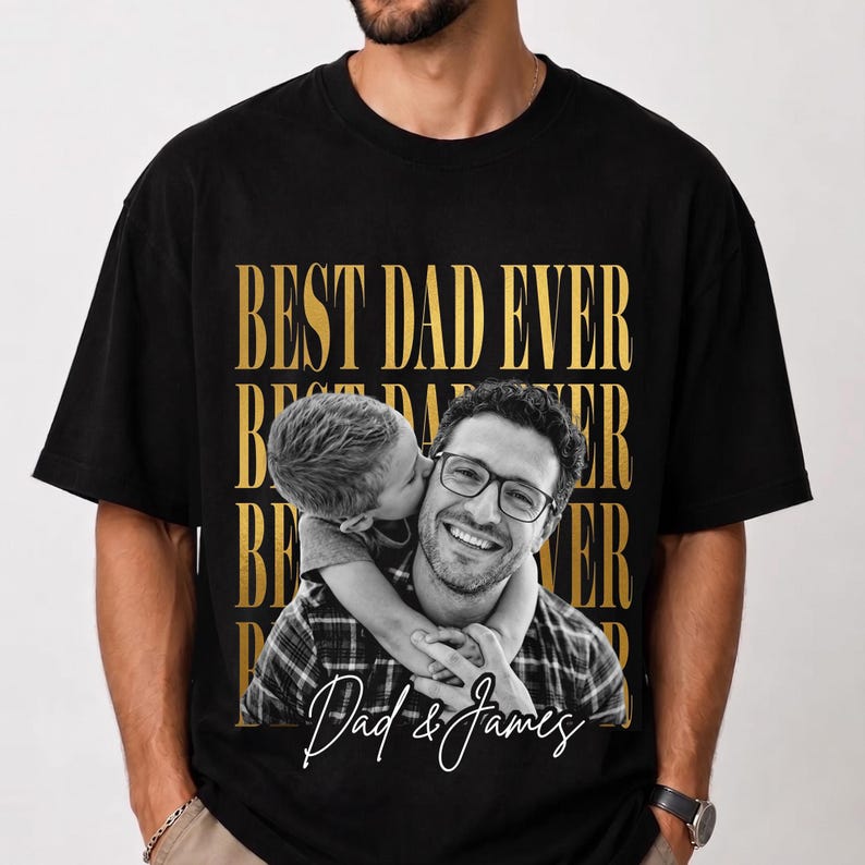 K&ouml;nnte beinhalten: Schwarzes T-Shirt mit goldenem Aufdruck "BEST DAD EVER". Das Shirt zeigt ein Schwarz-Wei&szlig;-Foto eines Mannes mit Brille, der von einem Kind gek&uuml;sst wird. Der Text "Dad & James" ist kursiv geschrieben.