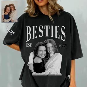 Peut inclure: T-shirt noir avec le mot "BESTIES" en blanc, avec "EST. 2016" en dessous. Une photo en noir et blanc de deux personnes souriantes qui s'embrassent est centrée sous le texte. Une petite photo des mêmes personnes est dans le coin supérieur gauche.