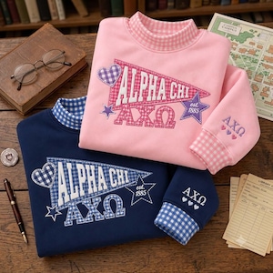 May include: A pink sweatshirt with a gingham mock neck and cuffs, featuring an embroidered pennant with the text Alpha Chi and AXO, plus a star and heart. A navy blue sweatshirt with a matching gingham mock neck and cuffs, featuring an embroidered pennant with the text Alpha Chi and AXO, a star, and a heart.