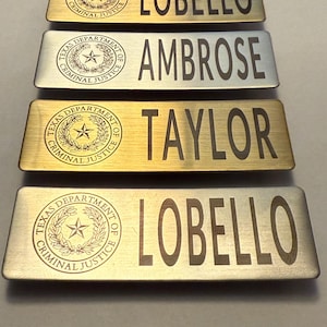 May include: Four rectangular name badges with the text "LOBELLO", "AMBROSE", "TAYLOR", and "LOBELLO". Each badge has the "TEXAS DEPARTMENT OF CRIMINAL JUSTICE" seal. The badges are in gold and silver tones.
