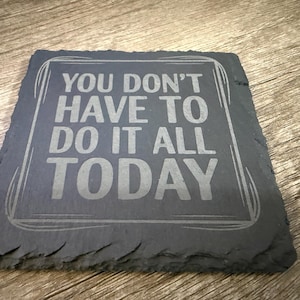 May include: A square, dark gray slate coaster with the engraved text "YOU DON'T HAVE TO DO IT ALL TODAY" in a light gray color. The coaster has a natural, rough edge and is placed on a wooden surface.