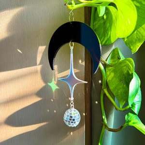 May include: A black crescent moon-shaped suncatcher with a star and disco ball pendant. The star has an iridescent finish, and the disco ball is made of small mirrored squares. The suncatcher hangs from a gold-colored chain.