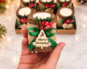 May include: A cylindrical candle holder with a bark-textured exterior wrapped in twine, featuring a green ribbon, red berries, and a wooden Christmas tree cutout with the text Merry Christmas. The candle is white and wax-filled.