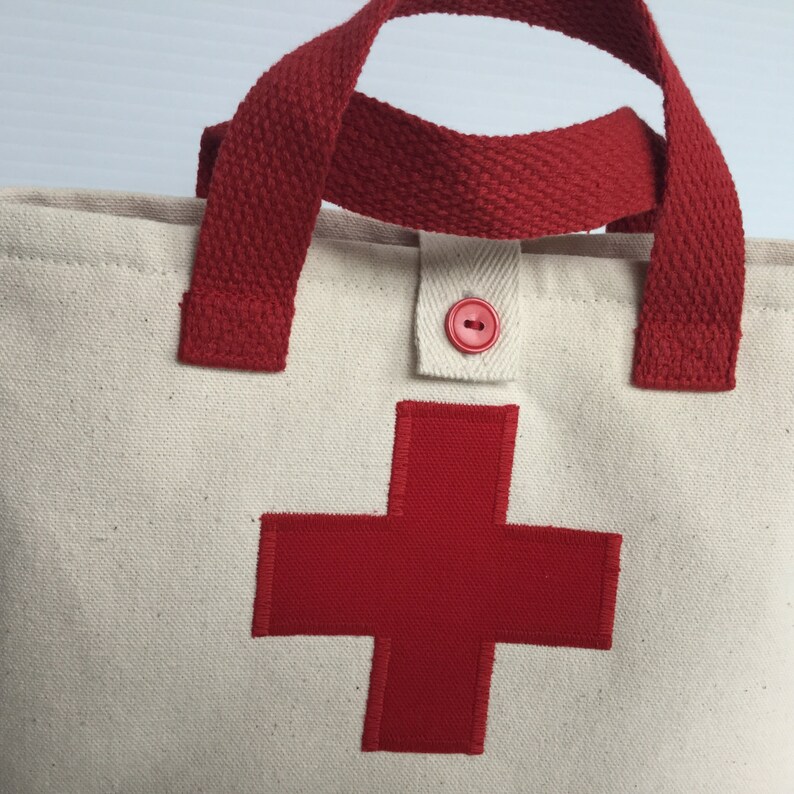 May include: Cream-colored canvas tote bag with a large red cross applique. Features red handles and a red button closure. The bag is designed for carrying items and has a simple, functional design.