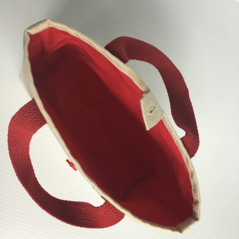May include: A small tote bag with a red interior and red handles. The exterior is a natural beige color. The bag has a button closure and is designed for carrying items. The bag is made of a sturdy material.