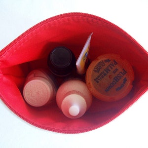 May include: A red zippered pouch with four small bottles and a round container inside. The bottles are clear and contain liquids. The round container is yellow and has blue text that reads "Noodhshed Palmwave"