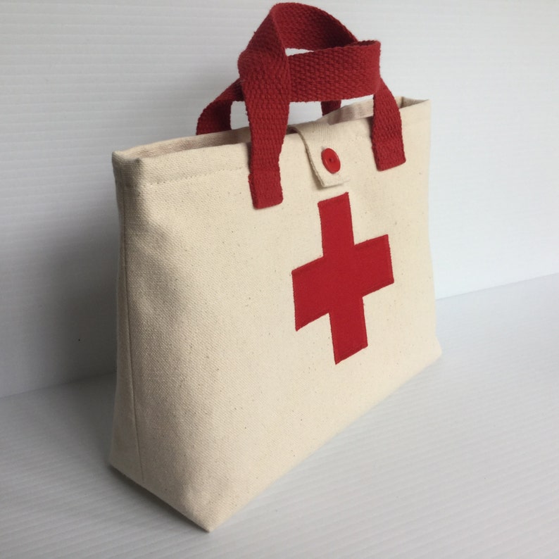May include: Cream-colored canvas tote bag with a large red cross emblem on the front. The bag has red handles and a red button closure. The bag is designed to carry medical supplies.