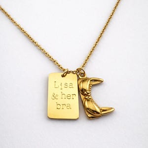 Könnte beinhalten: Goldfarbene Halskette mit zwei Anhängern. Ein Anhänger ist ein rechteckiges Etikett mit dem eingravierten Text "Lisa & her bra". Der andere Anhänger ist eine Krabbenschere. Die Halskette ist ebenfalls goldfarben.